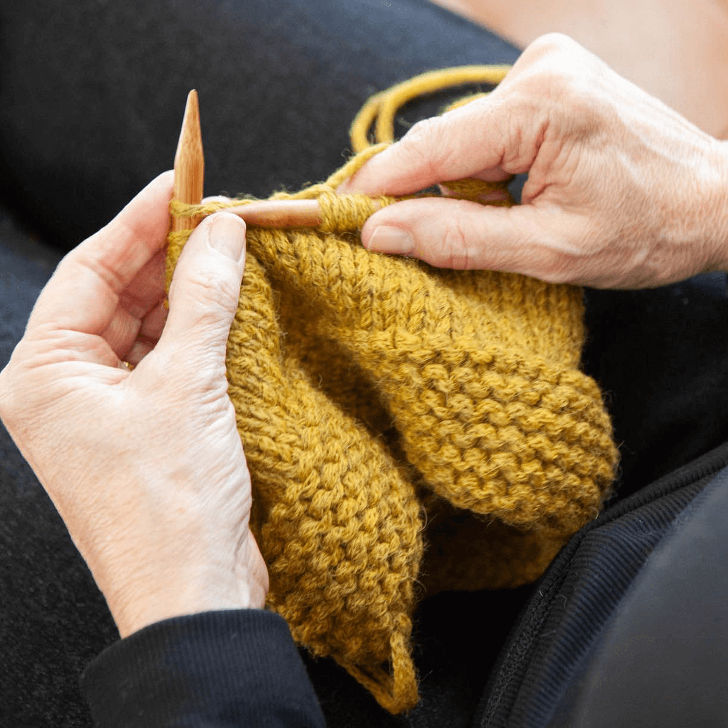 Hands knitting with mustard yellow yarn and wooden needles.